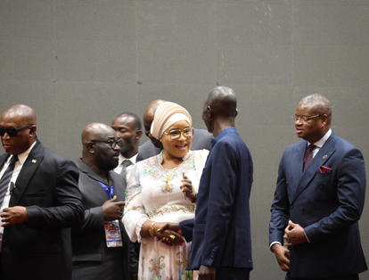 Group of formally dressed people on stage; a woman in a beige hijab chats with a man.
