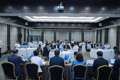 Large conference room with attendees around long tables and a presentation at the front.