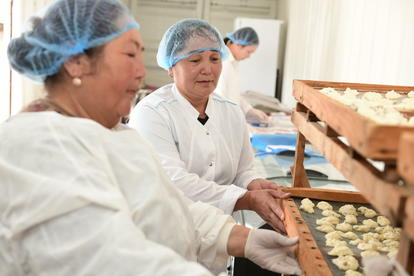 Two bakers in hair nets and white coats inspect dough rounds on a wooden rack.