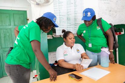 Three volunteers in green shirts and blue caps assist a woman at a registration desk.