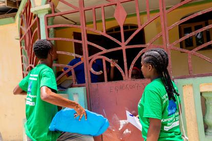 Photo of two students in green shirts beside a pink metal gate, one holding a blue folder.