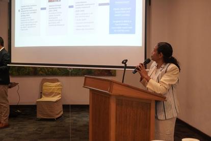 Woman at podium presenting with projection slides in a conference room.