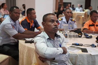 Man in light blue shirt sits with arms crossed at a conference table; others in uniforms nearby.