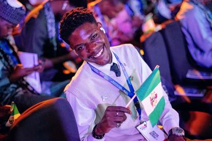 Smiling man in white shirt with lanyard holds Nigerian flag at a conference.