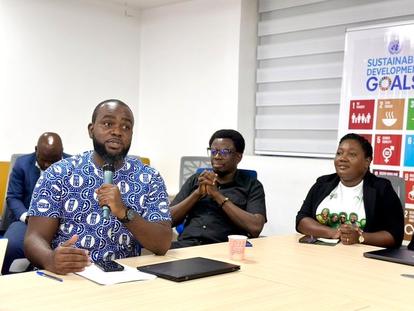 Photograph: three men at a table in a meeting; left man gestures, GOALS banner in the back.