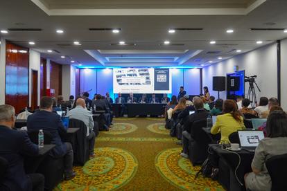Photograph of a conference: attendees seated at tables watching a blue-lit stage presentation.