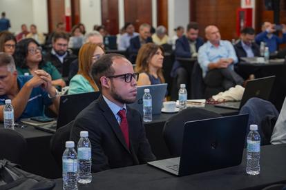 Attendee in suit with red tie at a table with laptop, conference crowd in background.