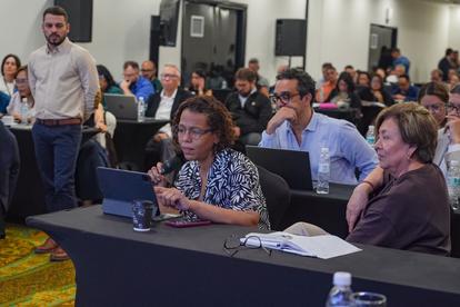 Panelists seated at a long table with laptops during a conference; audience visible in background.