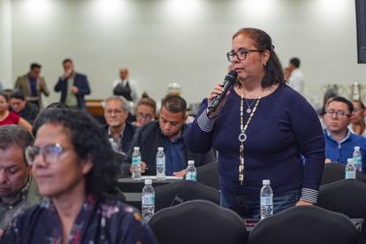 Woman with glasses in a blue top stands and speaks to a conference audience.