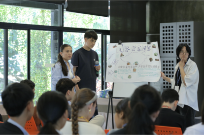 A presenter and two colleagues address a seated audience in a bright classroom with a whiteboard.