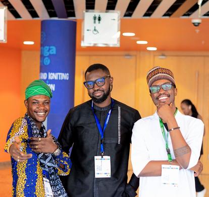Three people posing at a conference booth; left in colorful attire, middle in black, right in white.