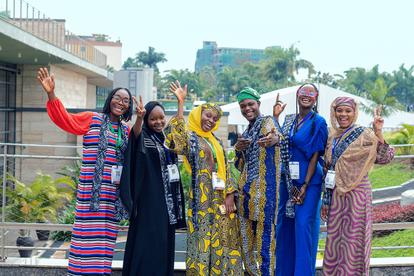 Group of seven people in colorful traditional African outfits posing on a sunny outdoor terrace.