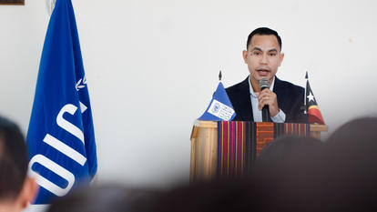 Man in a suit speaks at a podium with microphones, flanked by blue and dark flags.
