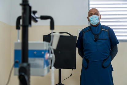 Healthcare worker in blue scrubs and mask with stethoscope beside medical equipment in a hospital.