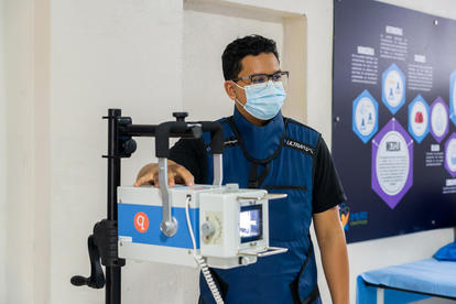Technician in blue uniform and mask demonstrates equipment on a stand, blue poster in background.