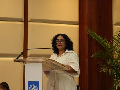 Woman in white blouse speaks at a lectern with UN emblem in a conference room.