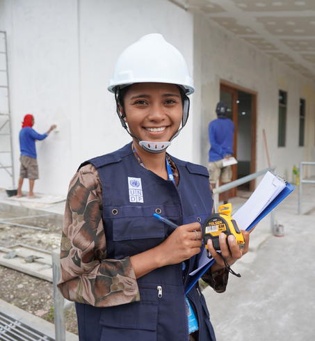 Construction worker in hard hat and safety vest holds clipboard near a building under construction.