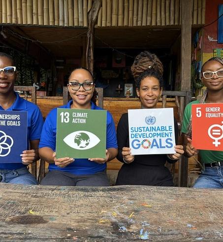 Four young women sit at a wooden bench outdoors, holding colorful SDG cards.