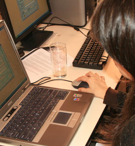 Woman with dark hair typing on a laptop at a cluttered desk with a second laptop.