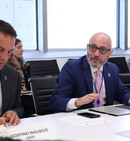 Three men in suits sit at a white conference table during a formal meeting; one writes notes.