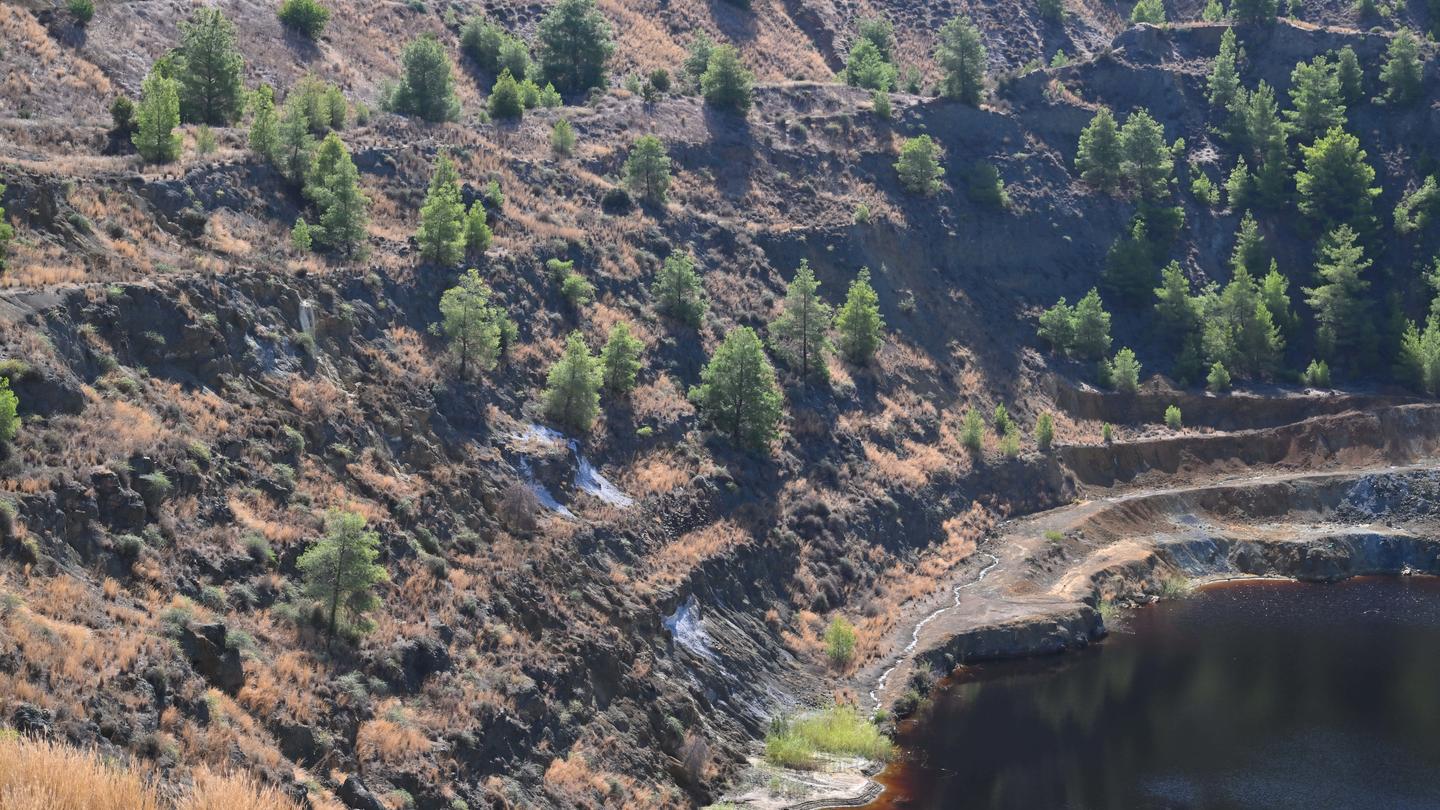 Steep rocky terrain with sparse trees surrounding a dark water-filled pit.