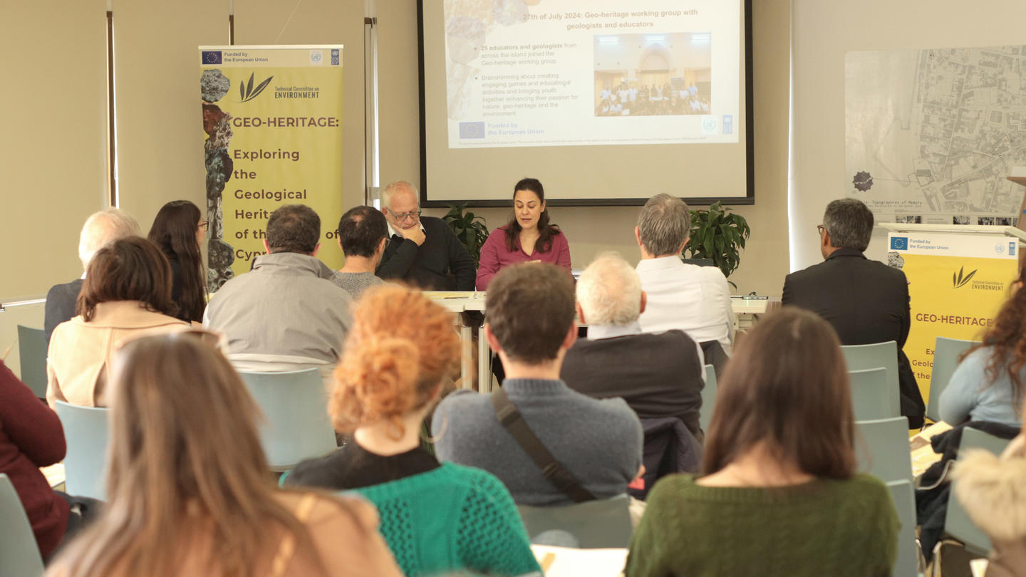 A conference scene with a speaker presenting to an audience seated in a bright room.