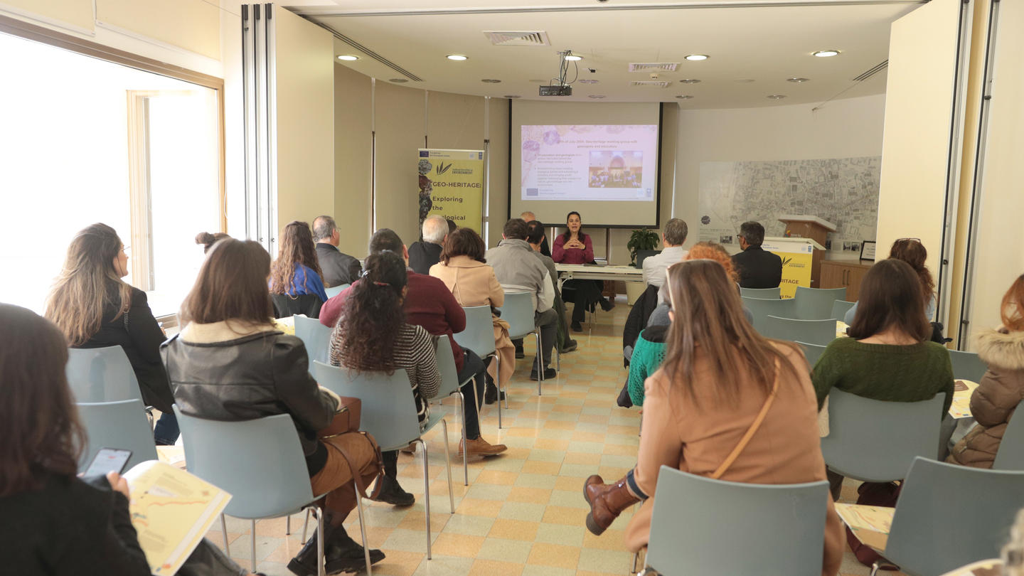 Audience seated in a conference room, facing a speaker and presentation screen.