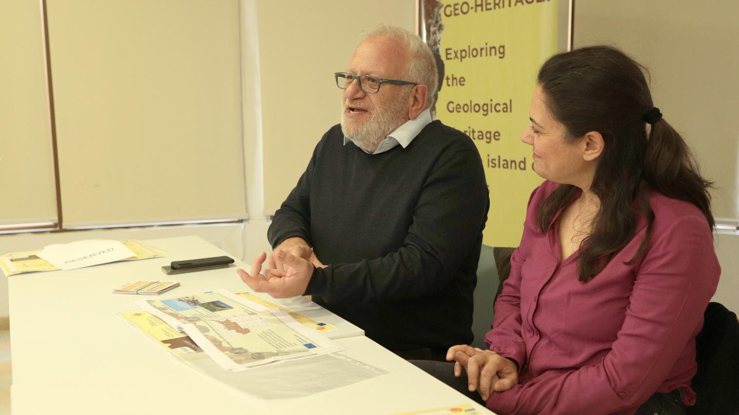 Two people seated at a table, engaged in conversation, with papers and a phone present.