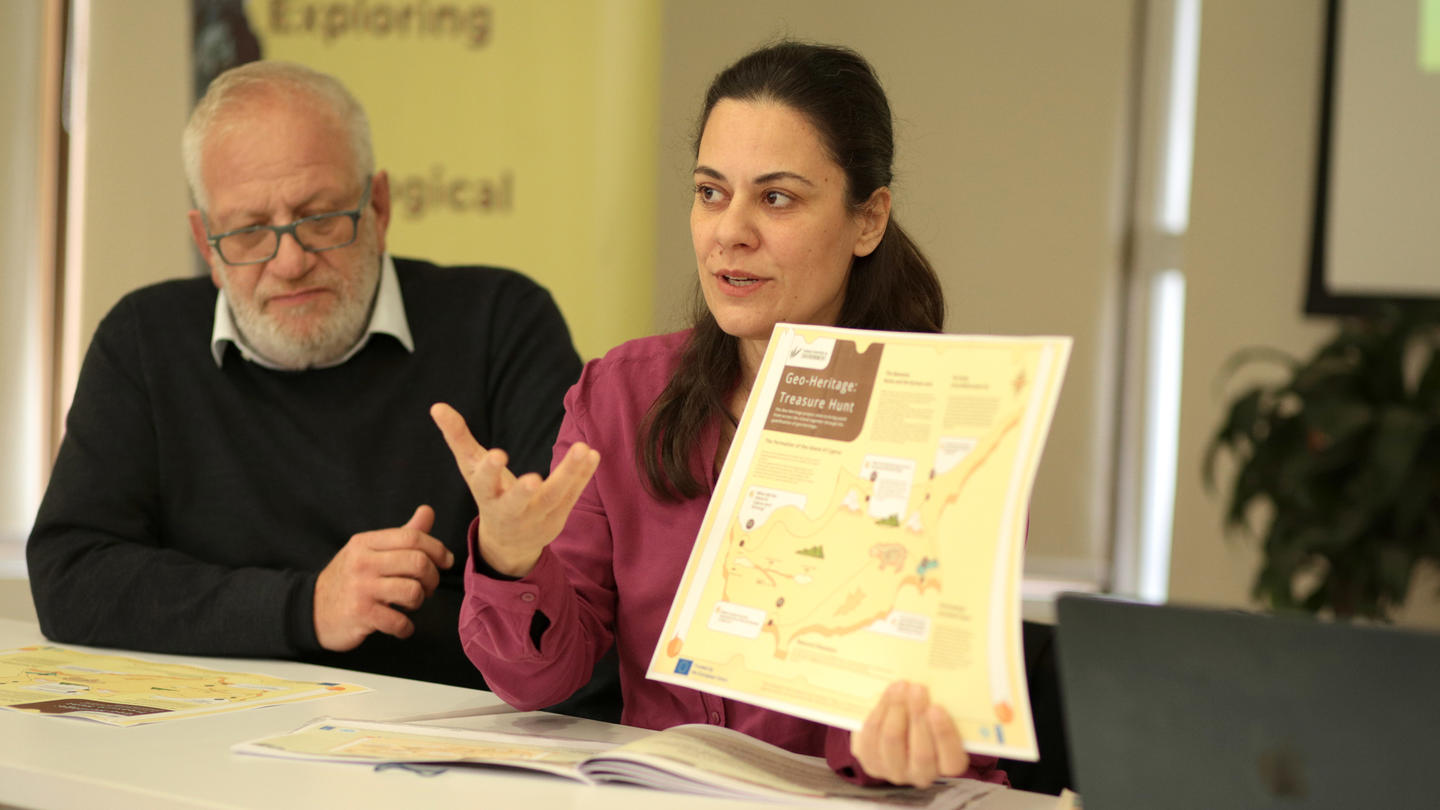 A woman in a pink blouse gestures while holding a map; a man listens nearby.