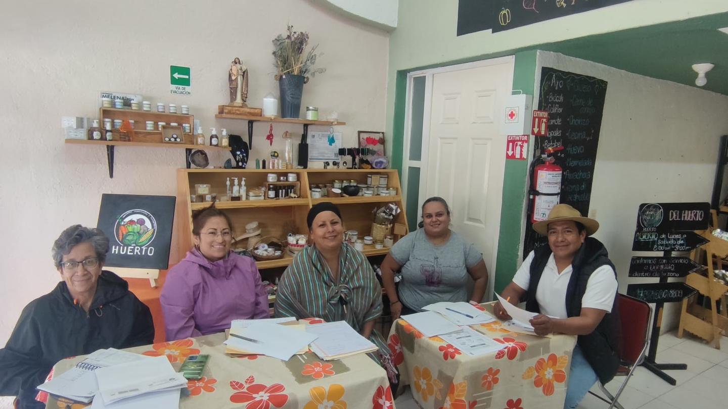 Five individuals sit around a table with floral patterns, discussing in a cozy interior setting.