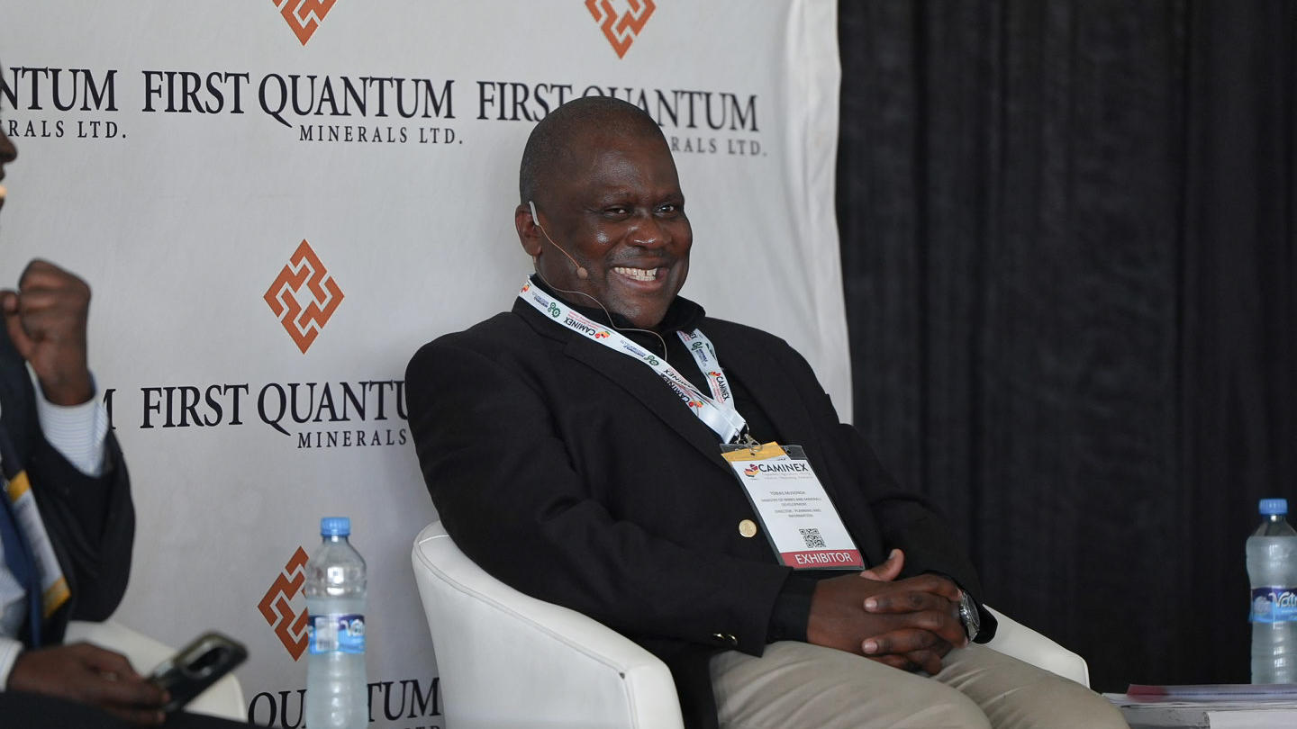 Smiling man in a suit seated on a panel, with a conference backdrop featuring logos.