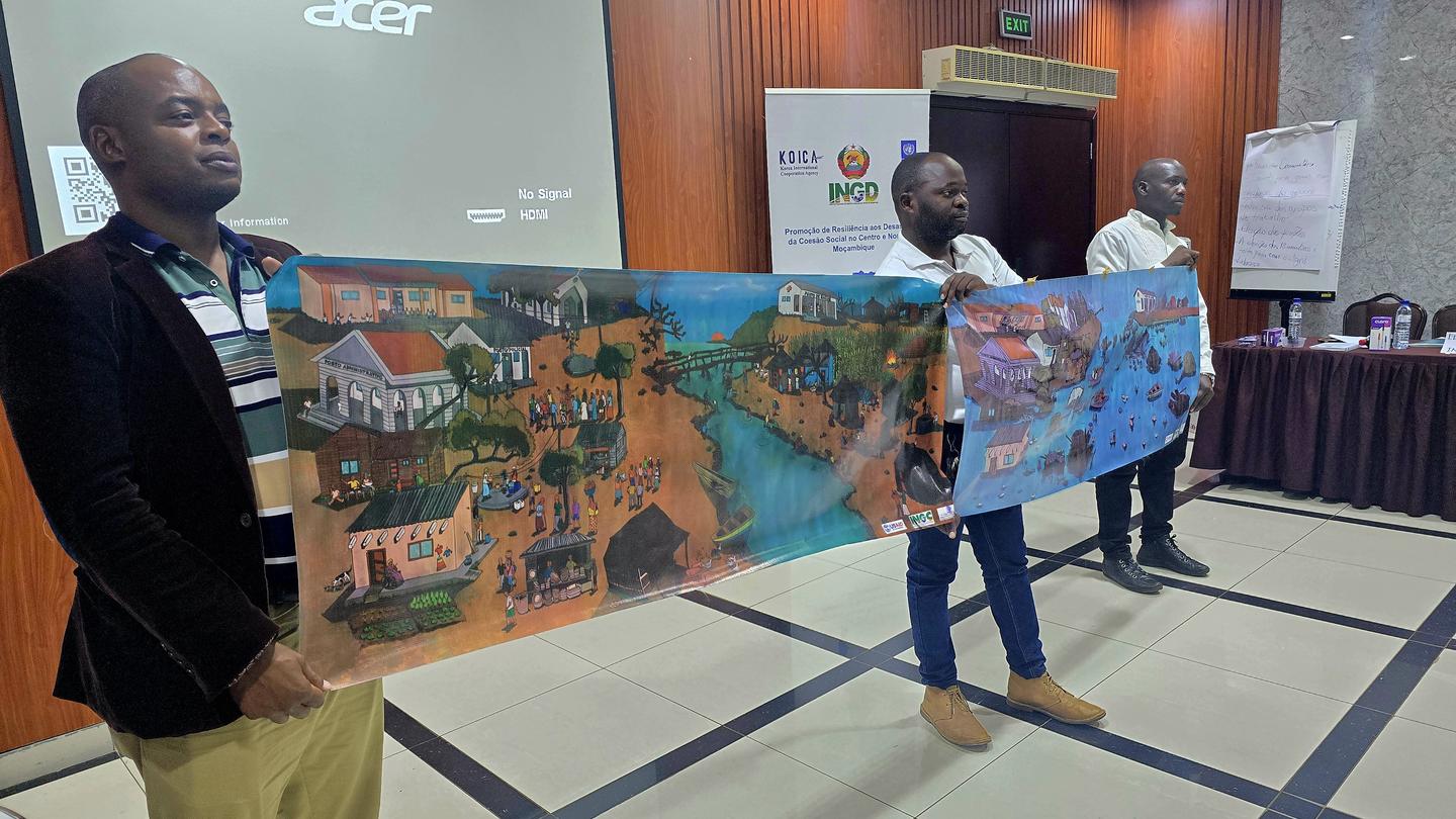 Three men hold large banners depicting vibrant scenes of a village at a conference room.