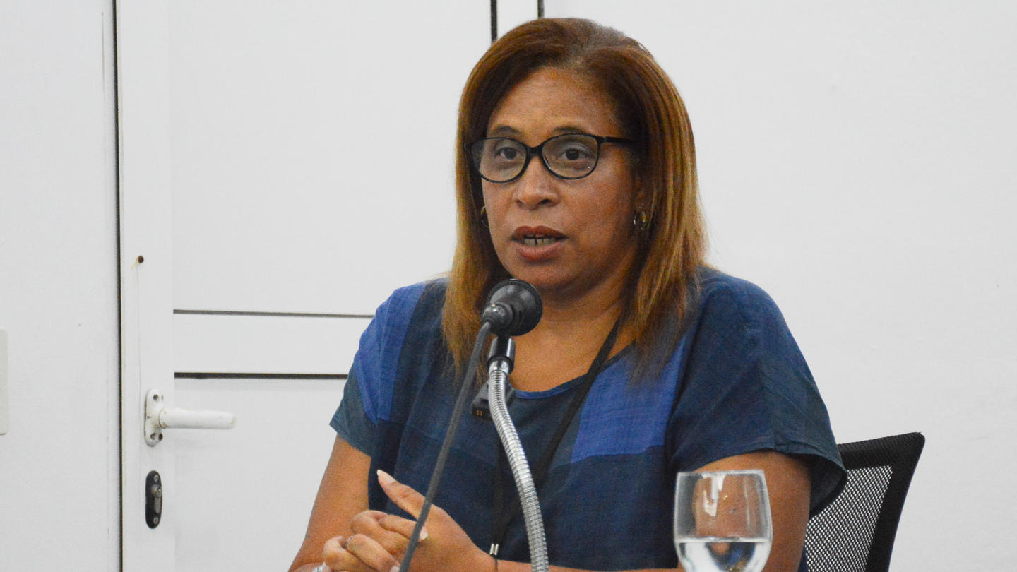 Person in blue shirt sits at a desk with a microphone and a water glass in a meeting room.