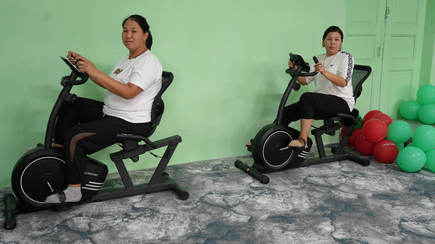 Two women on exercise bikes in a mint-green room with scattered balloons.