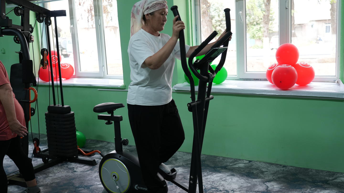 Photograph of a woman on a stationary bike in a bright gym, red balloons near a window.