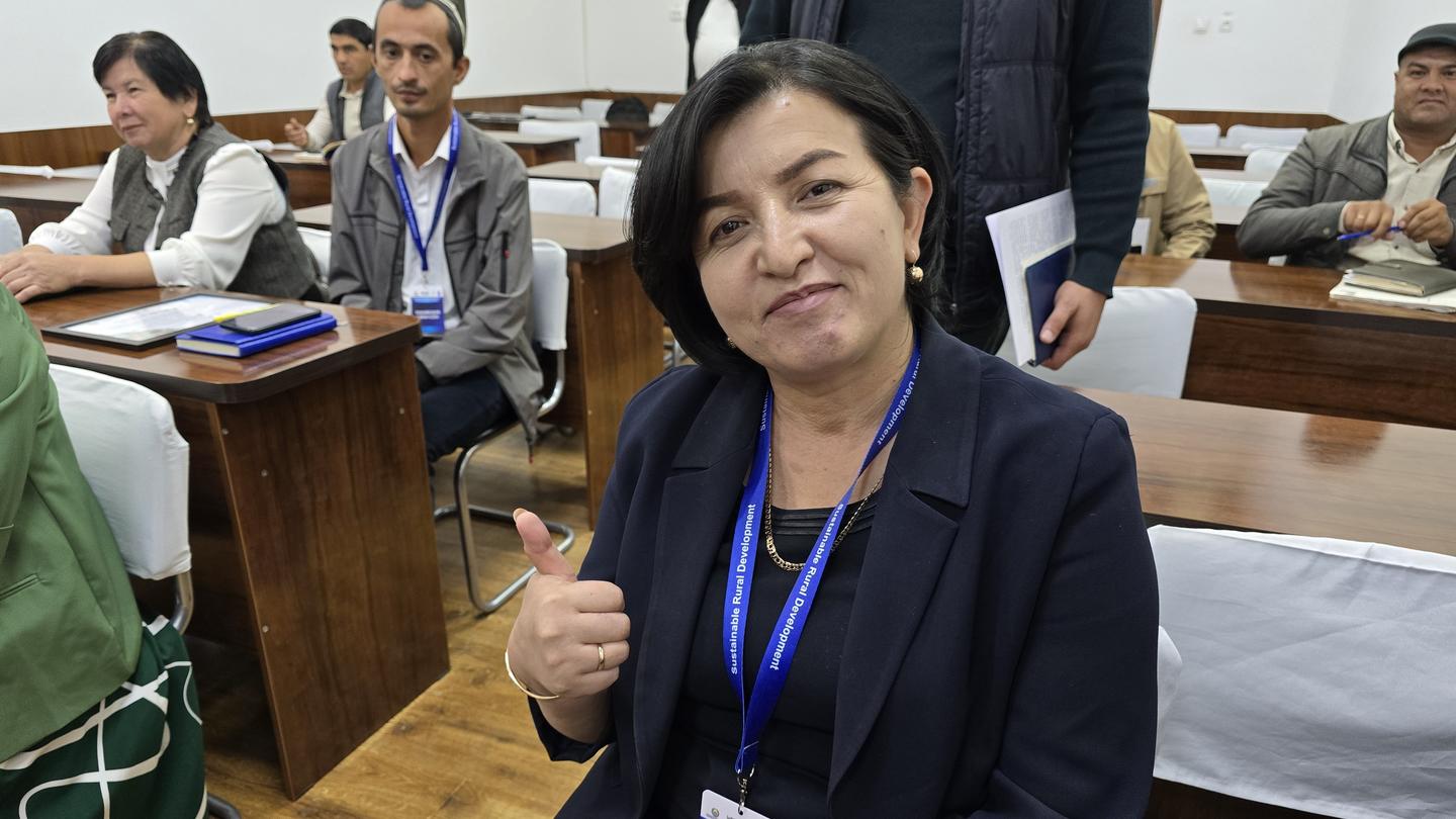 Person with a lanyard gives a thumbs up in a conference room; attendees sit at desks.