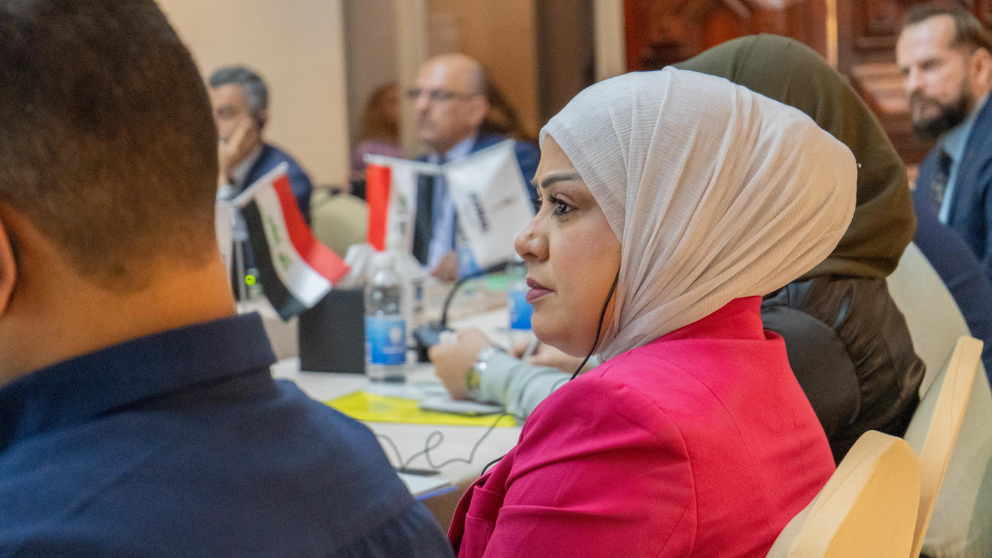 Participants at a conference table; foreground woman wearing beige hijab and pink top.