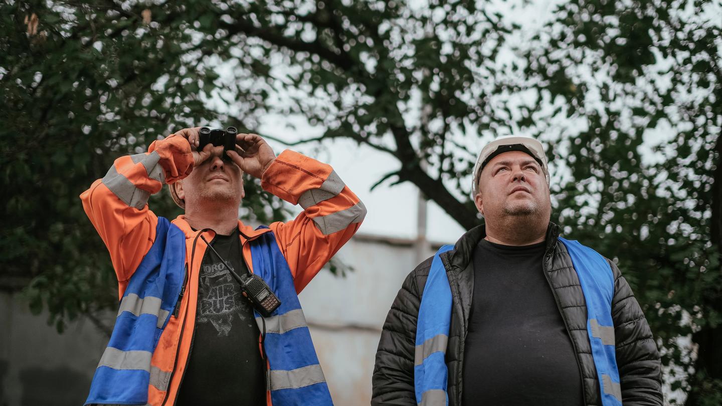 Two men outdoors looking up; one in orange jacket with blue safety vest, the other in a blue vest.