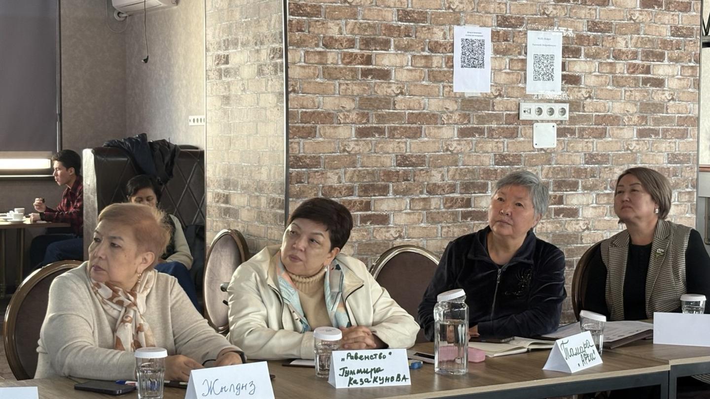 Five panelists sit at a table in a brick-walled meeting room.