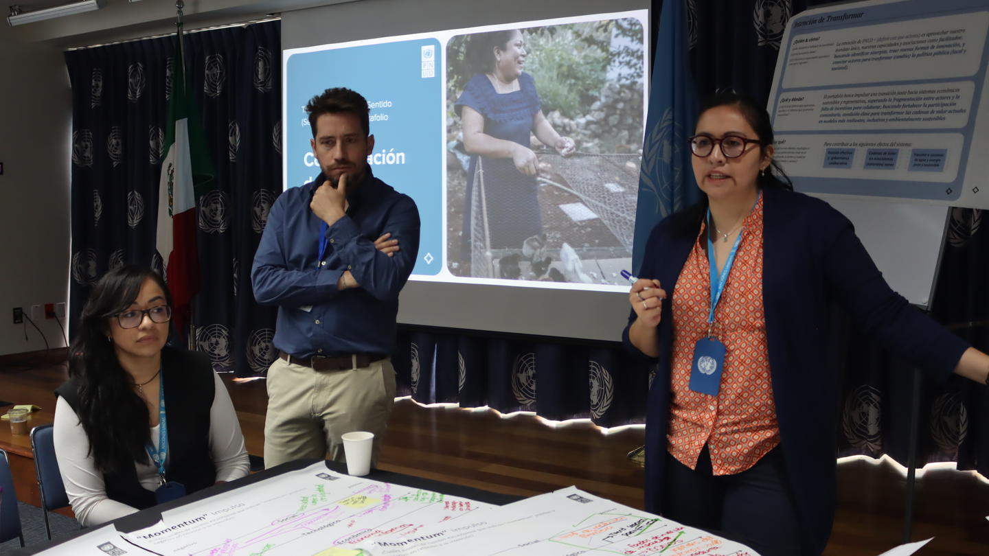 Group around a conference table with charts; man with microphone speaks, woman with lanyard stands.