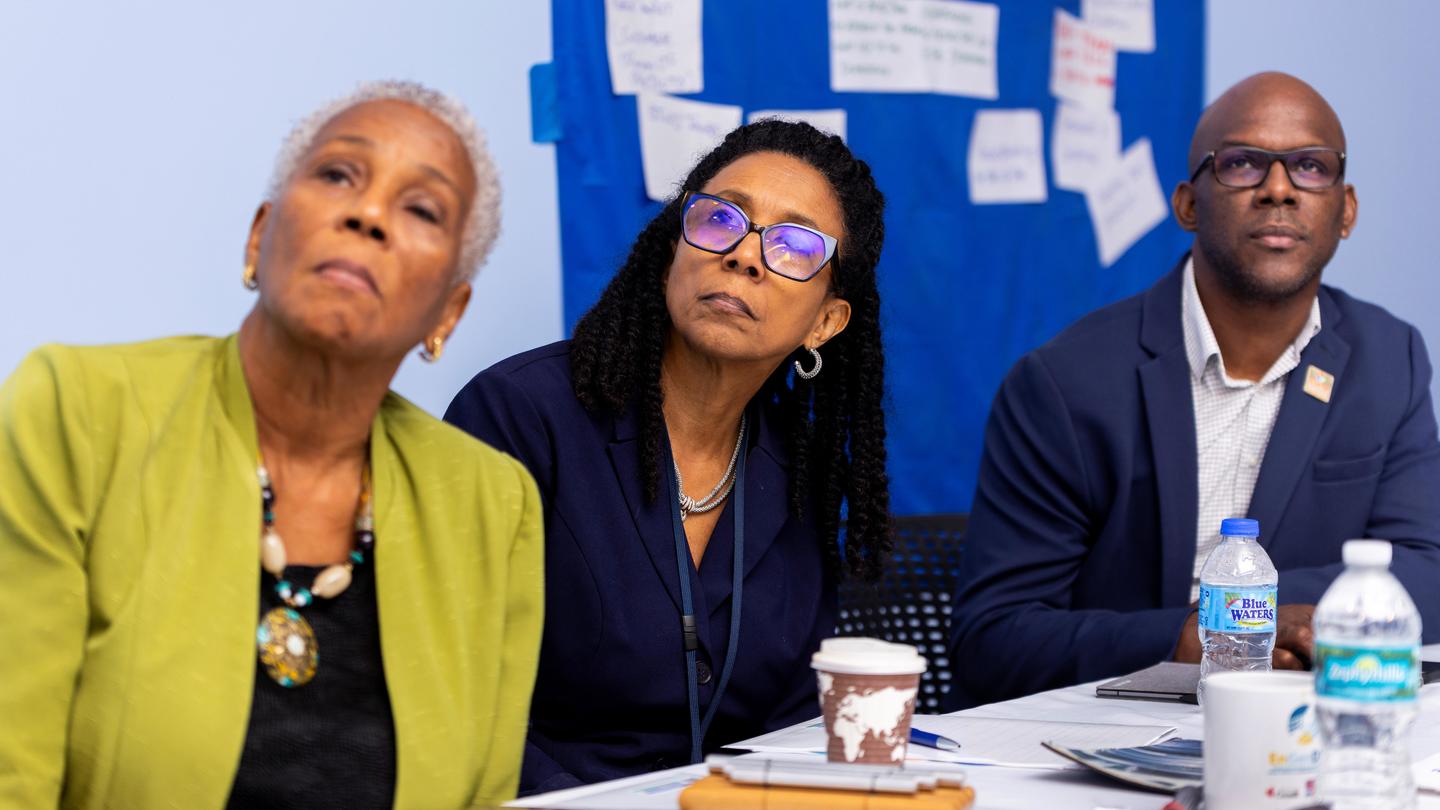 Photo: Three professionals at a table with papers and drinks, blue board with sticky notes behind.