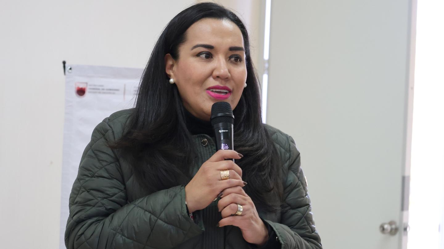 Person in a green quilted jacket speaks into a handheld microphone indoors.