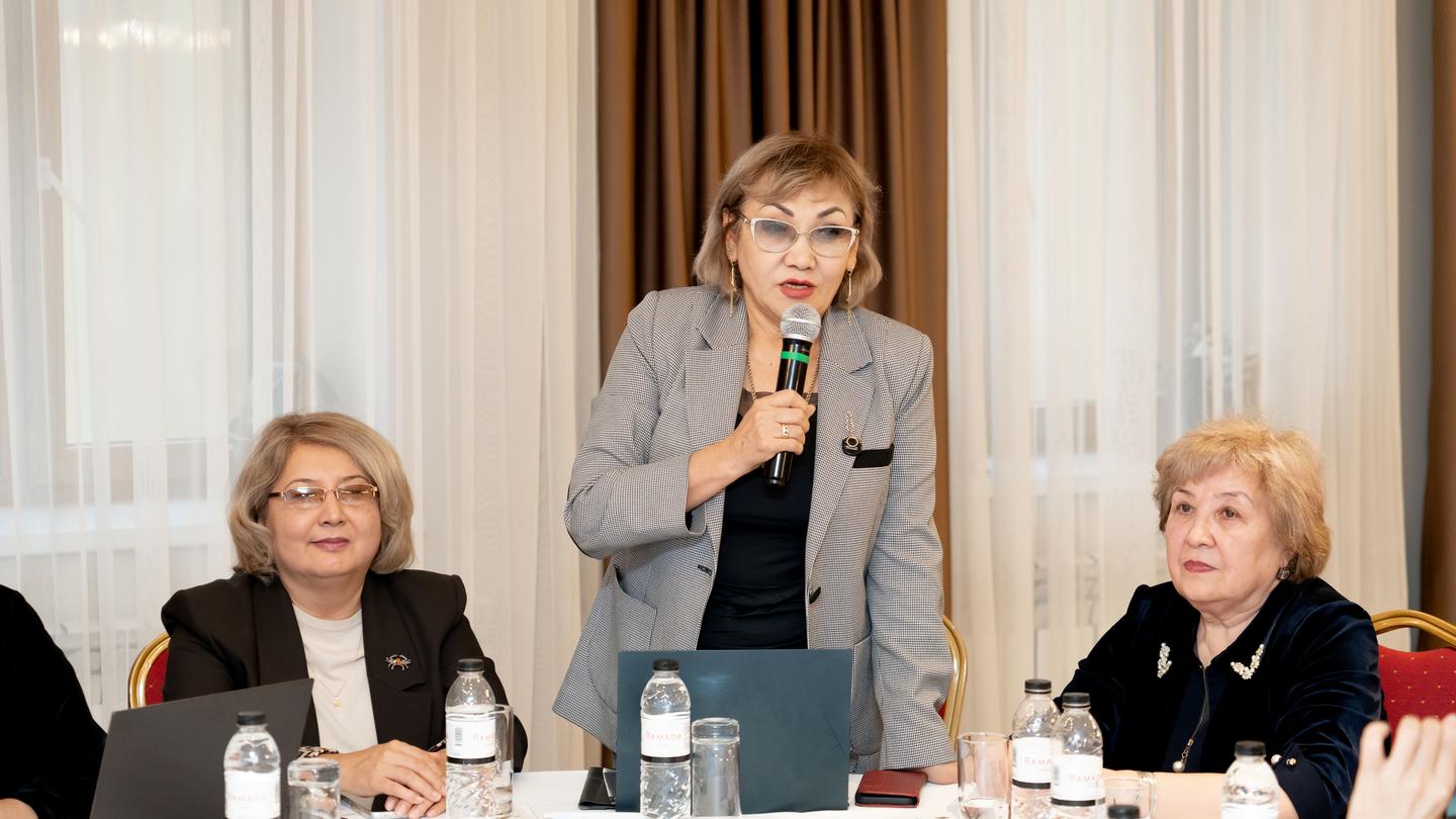 Photograph of a woman speaking into a microphone at a panel, with two seated colleagues.