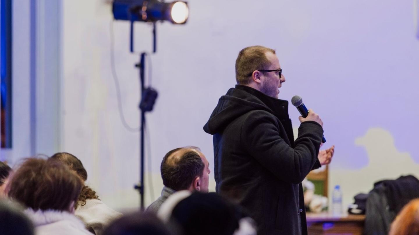 Photograph of a man in a dark jacket speaking into a microphone on stage, audience seated.