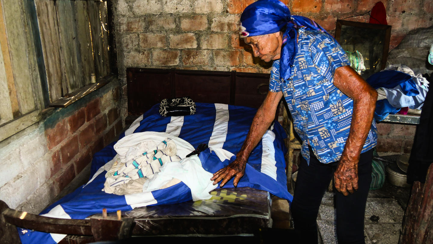 Man in a blue shirt and cap leans over a bed in a brick-walled room; body wrapped in white cloth lying on it.