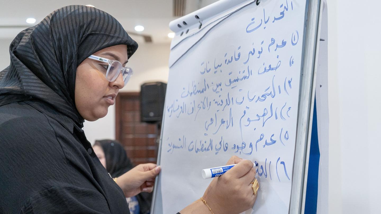 Photograph of a woman in a hijab writing on a vertical banner with blue script.