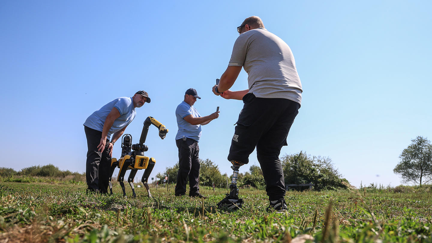 A robot and several people, including a veteran with an amputated leg, are in a field taking pictures
