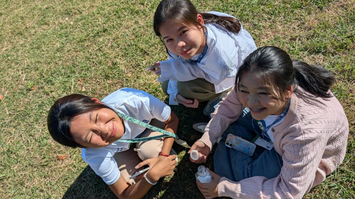 Three people sit on grass outdoors, looking up at the camera and smiling.