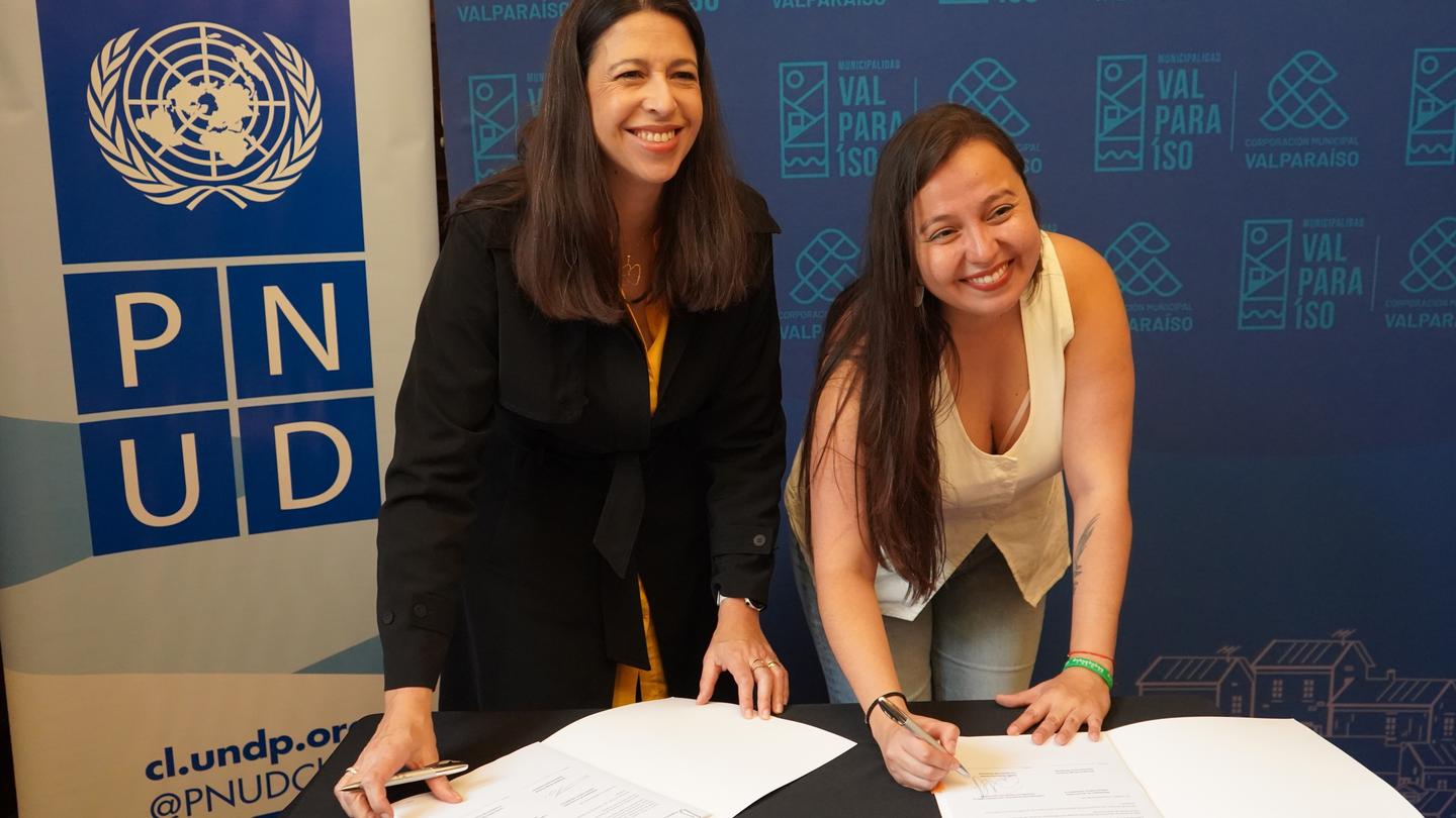 Two people signing a large guest book at a table, in front of a blue United Nations backdrop.