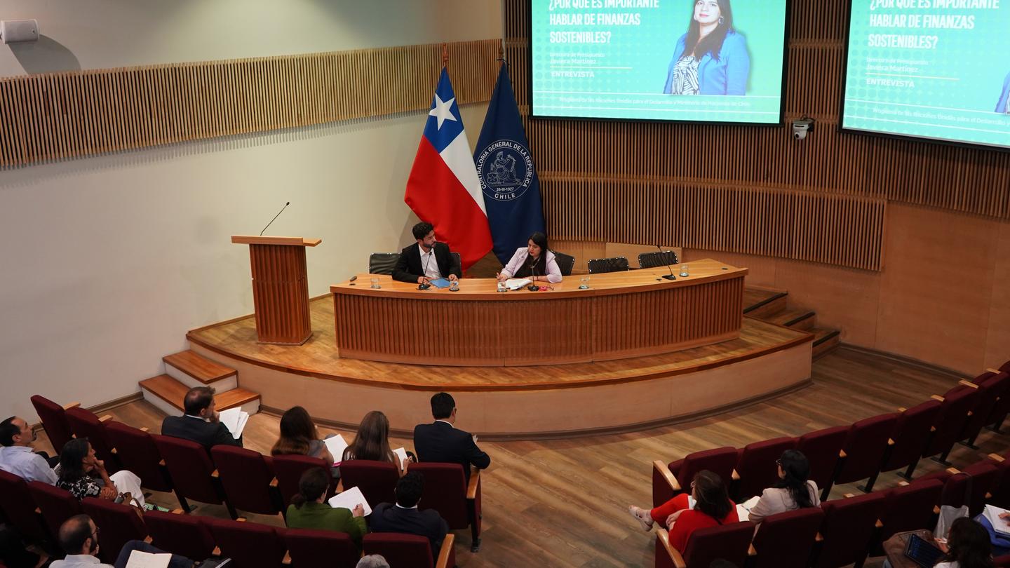Photograph of a speaker at a podium on a curved wooden stage, flags behind, audience listening, slides on screens.